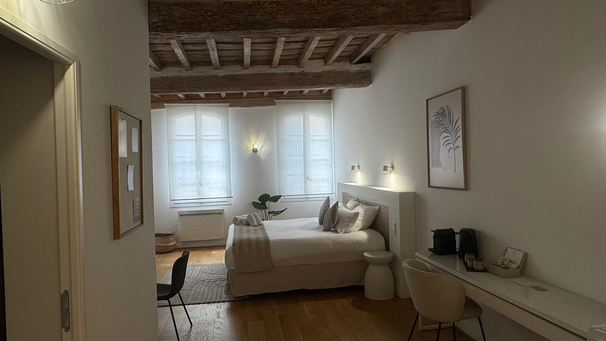Un dormitorio con cama, mesita de noche, silla y escritorio con otra silla. Hay ventanas con cortinas, un radiador y una planta.