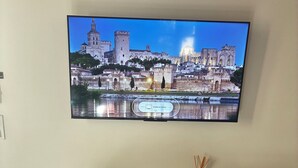Et fladskærms-tv på væggen viser et bybillede med et slot og en flod. Under tv'et er et træskrivebord med en stol, en fjernbetjening og nogle brochurer.