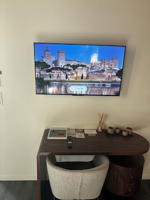 A flat-screen TV mounted on a wall displaying a cityscape with a castle and a river. Below the TV, there's a wooden desk with a chair, a remote control, and some brochures.