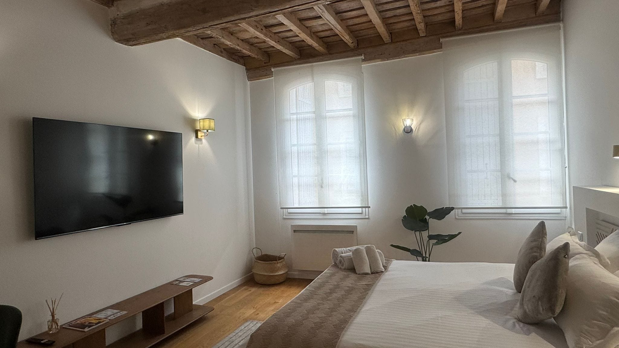 A bedroom with a large bed, a flat-screen TV mounted on the wall, and two windows with light fixtures.