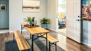 Uma área de refeições com mesa e bancos de madeira, plantas em vasos e uma porta que dá para o exterior.