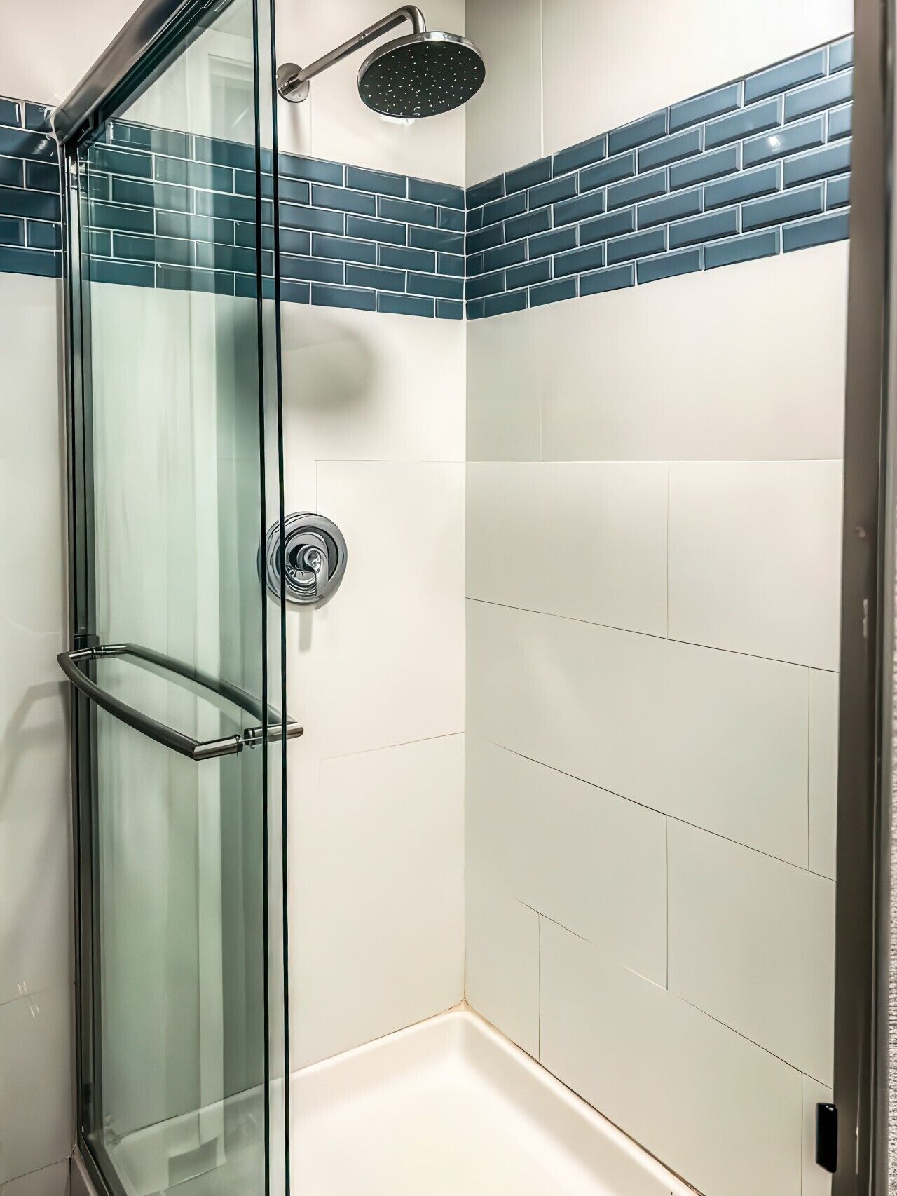 A glass-enclosed shower with a handheld showerhead, a glass door with a towel rack, and a corner wall with blue and white tiles.