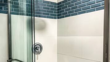 A glass-enclosed shower with a handheld showerhead, a glass door with a towel rack, and a corner wall with blue and white tiles.
