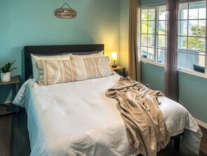 A neatly made bed with a white comforter and pillows, a bedside lamp, a small plant, and a window with curtains.
