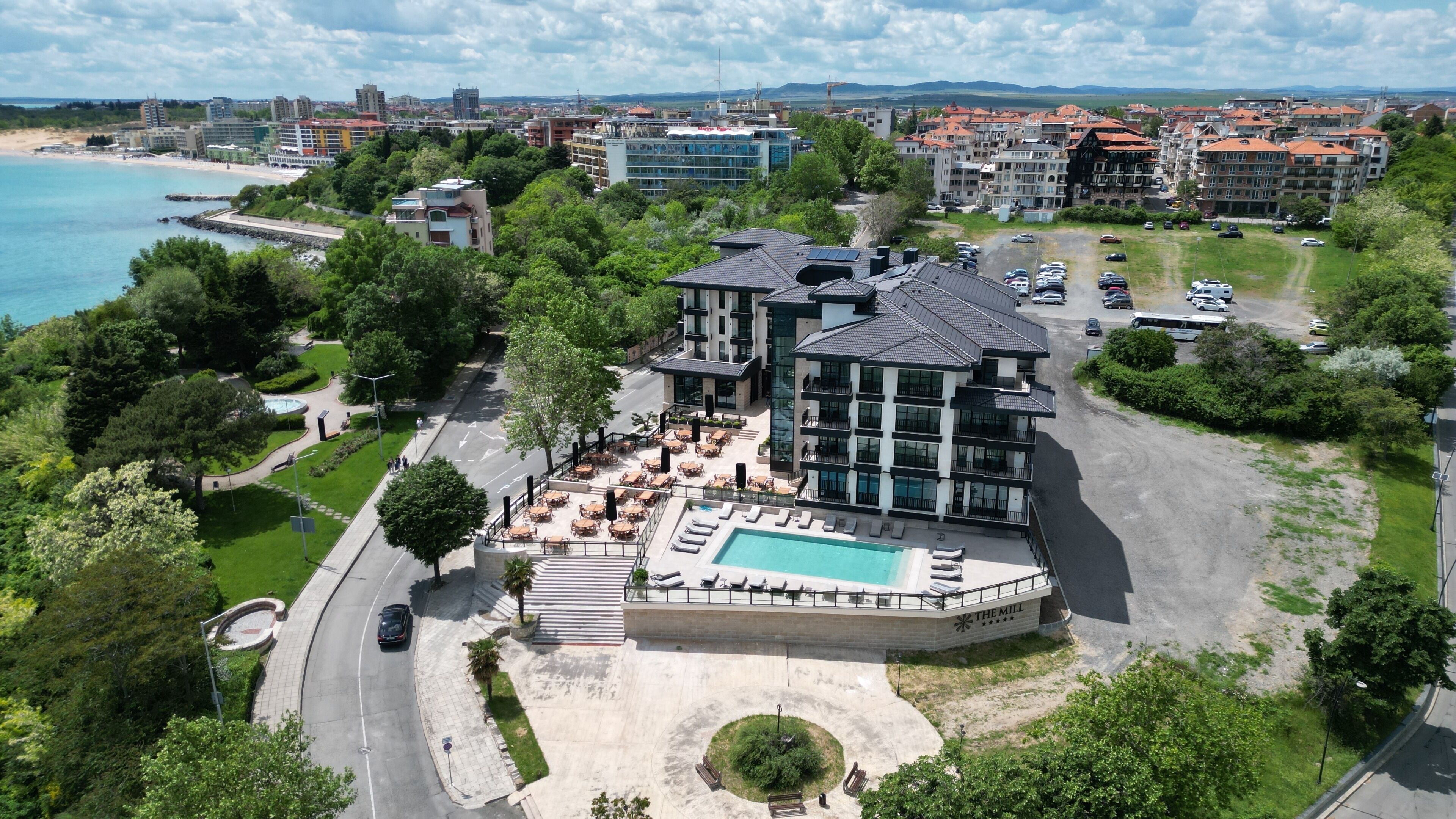 A multi-story hotel with a rooftop pool, surrounded by greenery and a view of the sea.