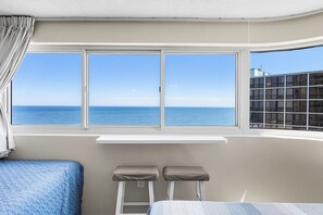 Ein Zimmer mit großem Fenster, einem Bett, zwei Hocker und einem weißen Regal.