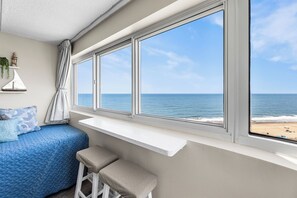 Une chambre avec une grande fenêtre, un lit avec une literie bleue, un petit tabouret et une étagère ornée d’objets décoratifs.
