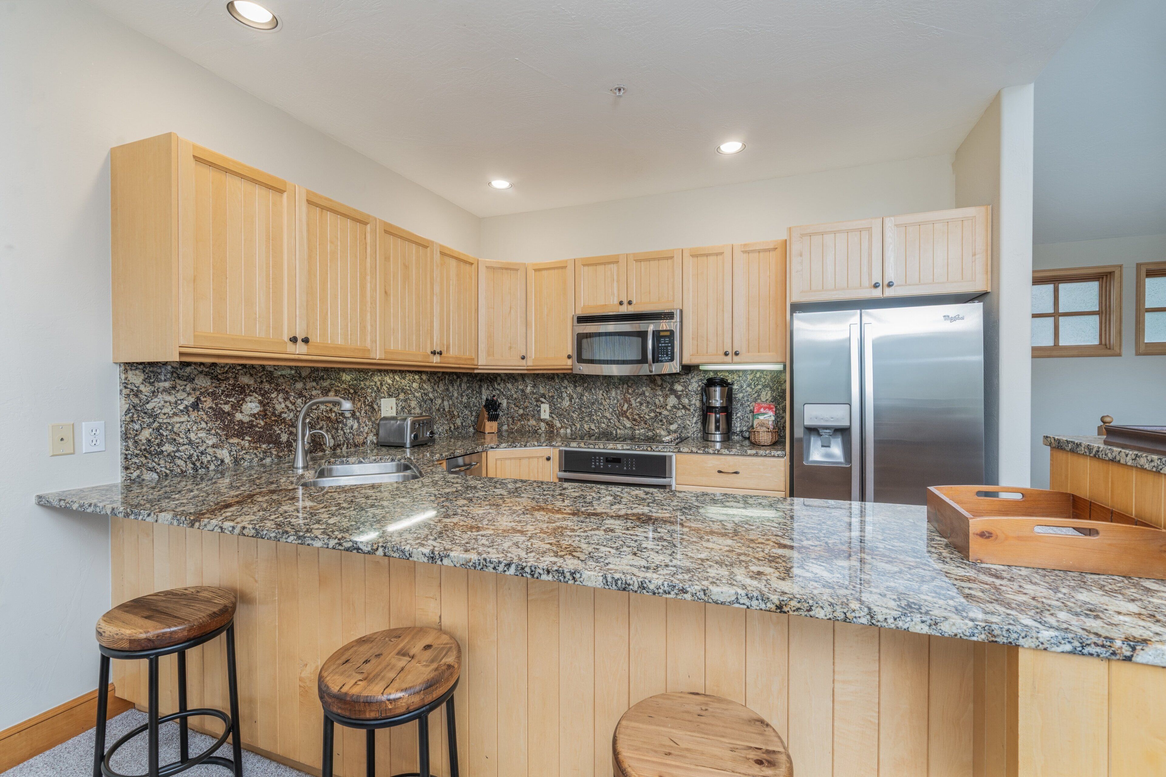 A kitchen with wooden cabinets, granite countertops, a stainless steel refrigerator, and two stools.