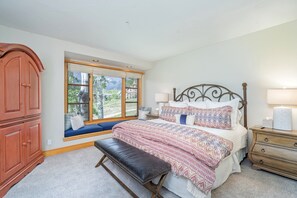 Un dormitorio con un ventanal grande, una cama con ropa de cama estampada, una banca, un aparador de madera y una mesita de noche con una lámpara.