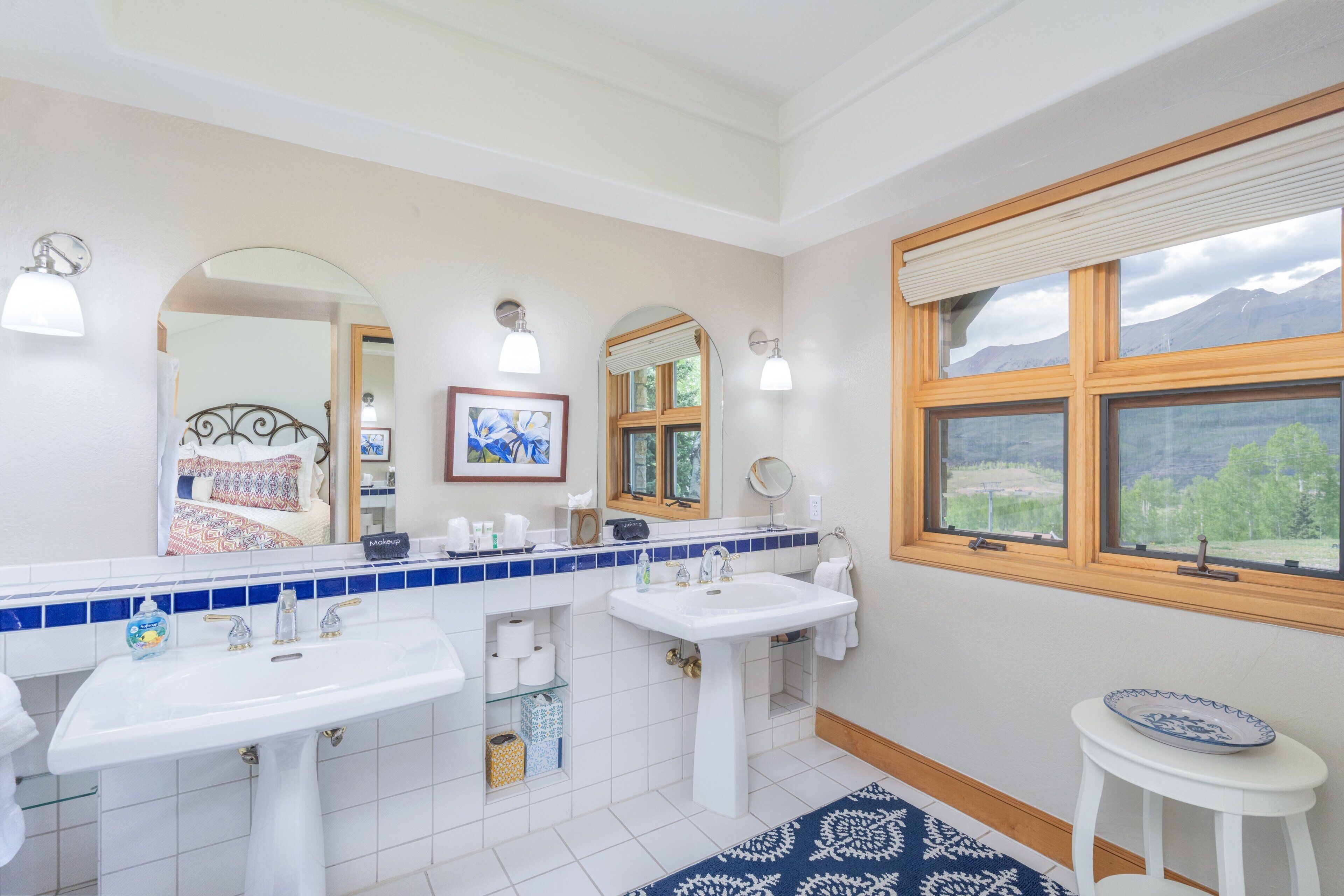 A bathroom with two sinks, a mirror, a window with a view, and a blue patterned rug.