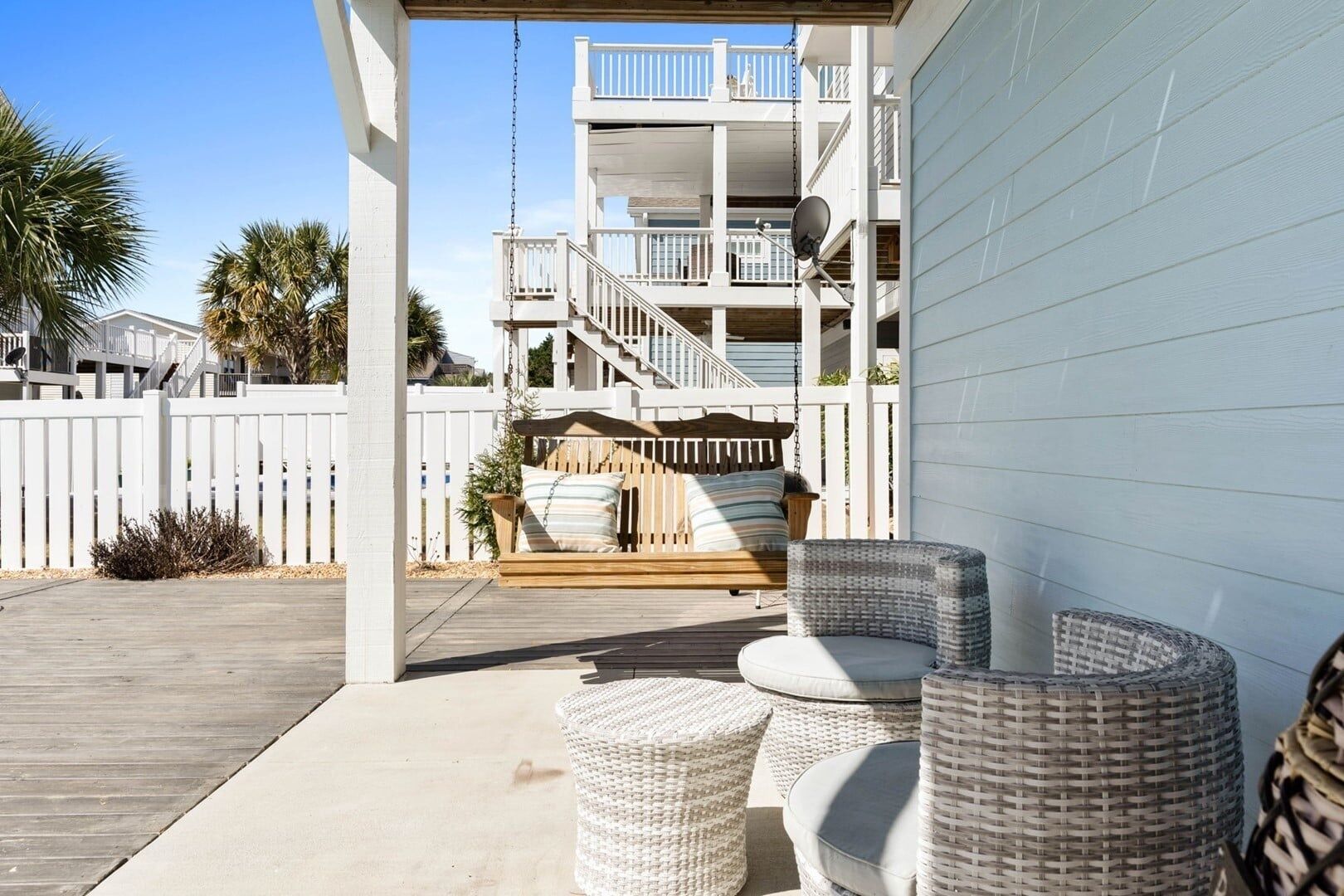 A covered outdoor seating area with wicker furniture, a wooden bench, and a white fence.