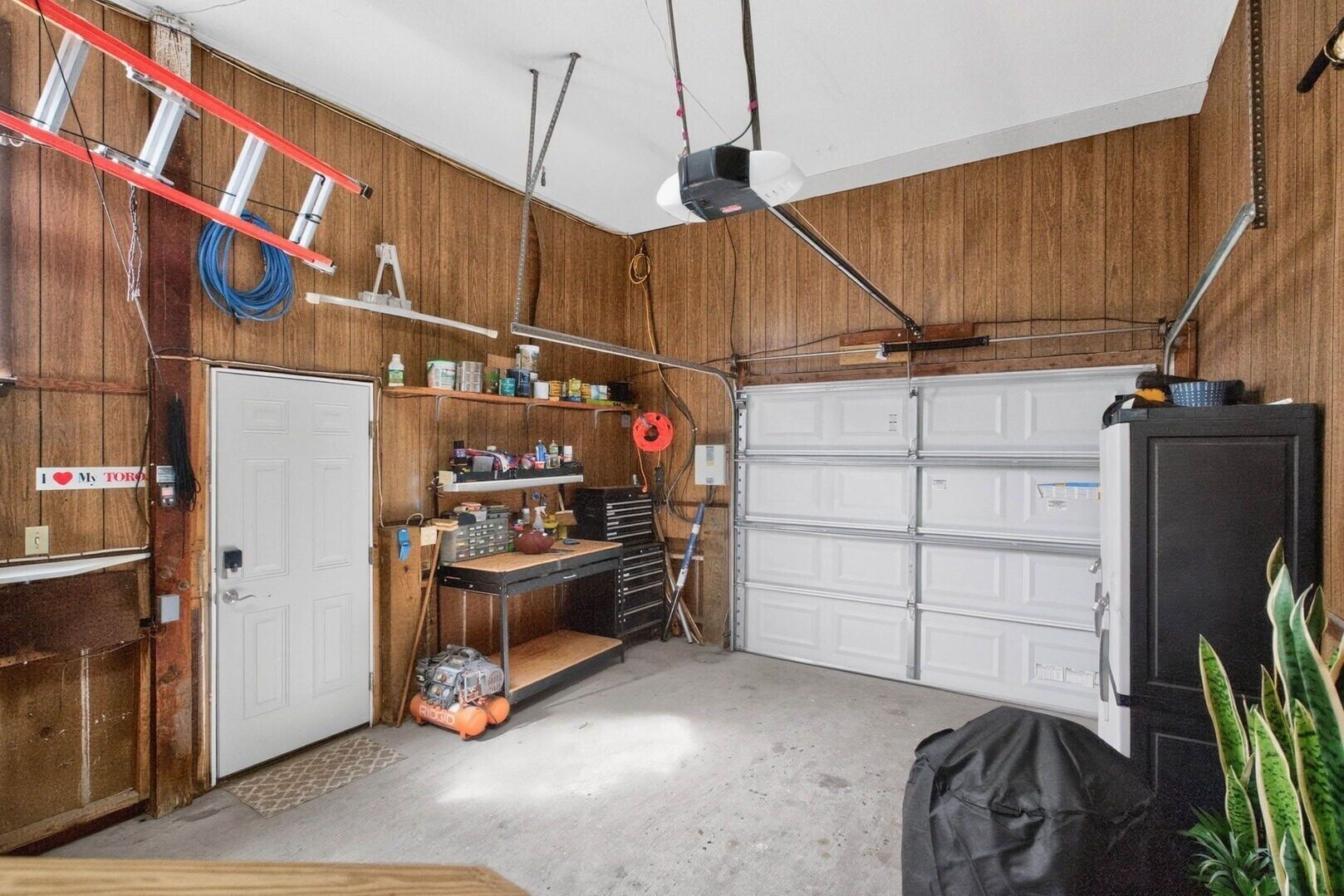 Un garage comprenant un établi, des étagères remplies d’outils, une échelle et une porte de garage.
