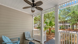 A covered porch with a ceiling fan, two Adirondack chairs, a patterned rug, and a view of a landscaped area with trees and a building in the background. - 30a Getaway 3 Bedroom Condo (Santa Rosa Beach)