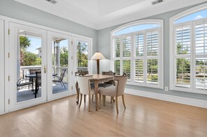 A room with a dining table, chairs, a lamp, and large windows with shutters.