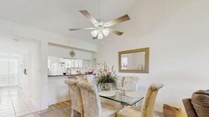 A dining area with a glass table, four chairs, a ceiling fan, a kitchen in the background, and a mirror on the wall. - Venice Condo, Pool, Balcony & Beaches Nearby! (Venice)
