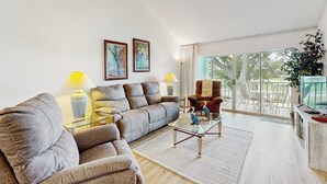 A living room with a sofa, armchair, glass coffee table, and a TV. There are two lamps, a plant, and a painting on the wall. - Venice Condo, Pool, Balcony & Beaches Nearby! (Venice)