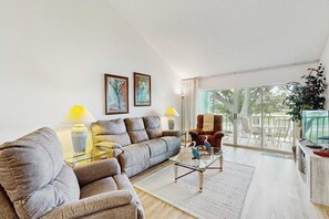 Ein Wohnzimmer mit einer Couch, einem Sessel, einem Glastisch und einem Fernseher. Zwei Lampen, eine Pflanze und ein Gemälde zieren die Wand.