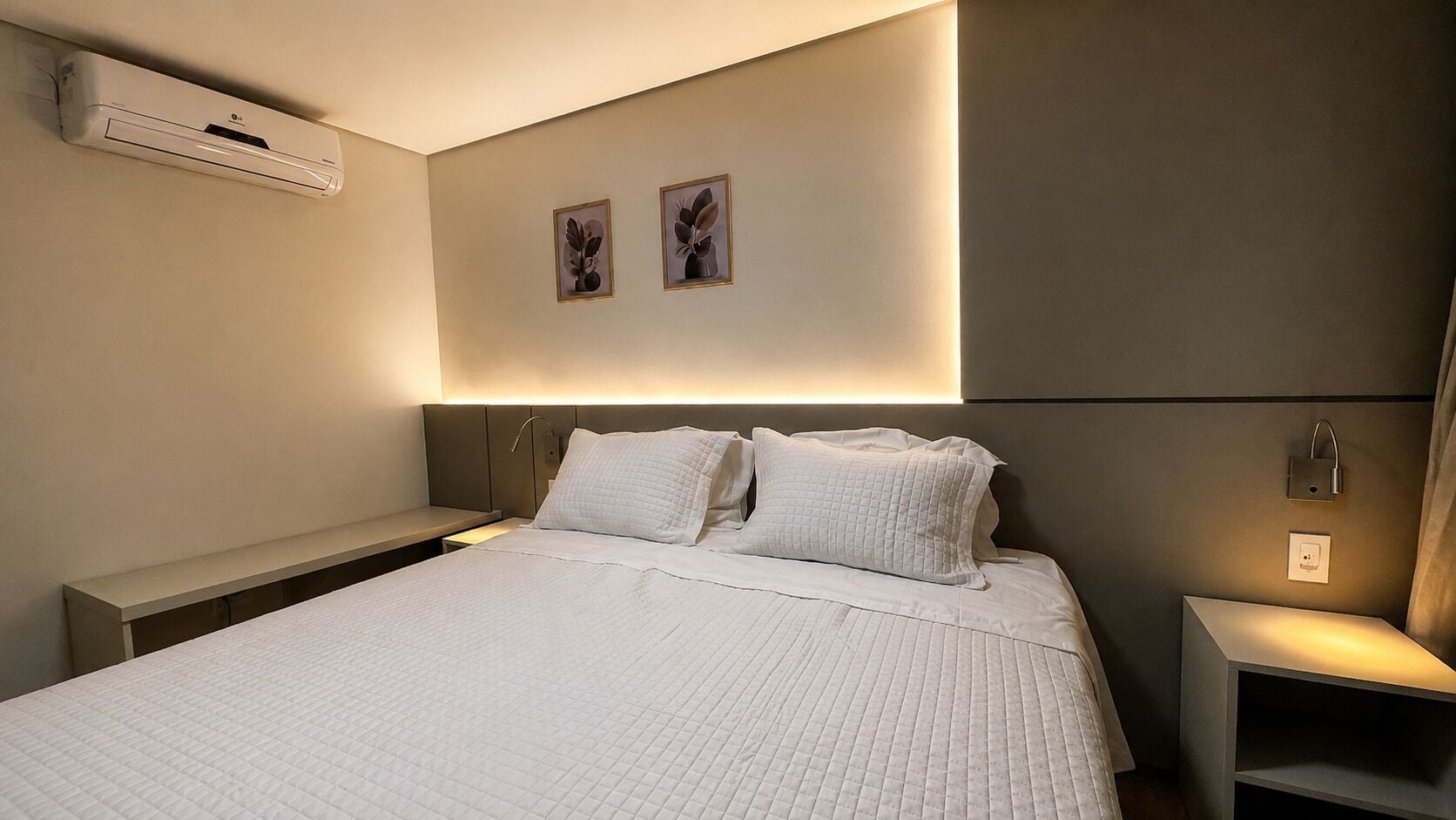 Una habitación de hotel moderna con una cama grande, mesitas de noche, un equipo de aire acondicionado, una obra de arte en la pared y una mesita de noche con lámpara.