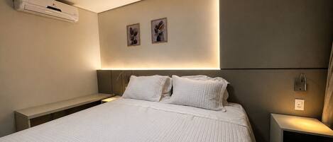 Una habitación de hotel moderna con una cama grande, mesitas de noche, un equipo de aire acondicionado, una obra de arte en la pared y una mesita de noche con lámpara.