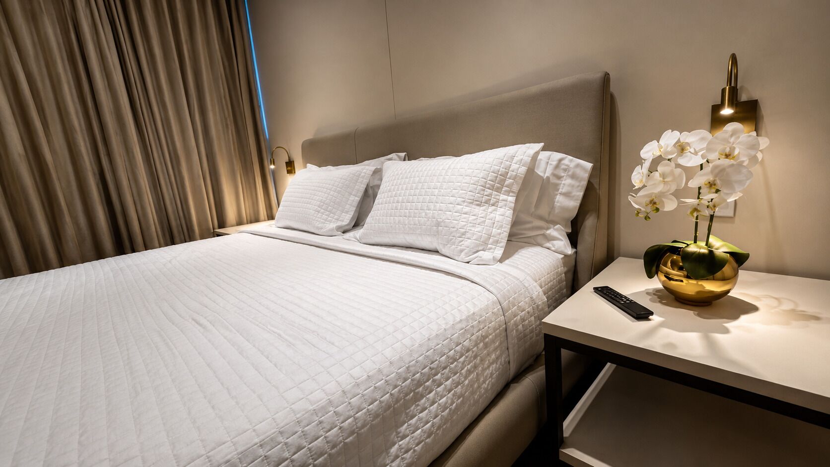 A neatly made bed with white bedding, a bedside table with a remote control, and a decorative orchid vase.