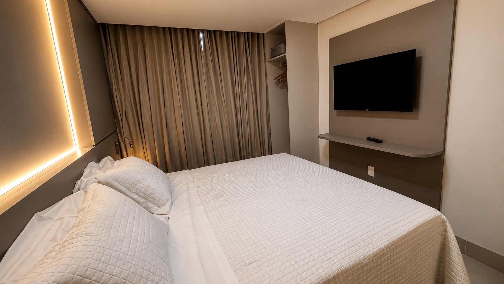 A modern hotel room with a neatly made bed, a flat-screen TV mounted on the wall, and a built-in shelf.