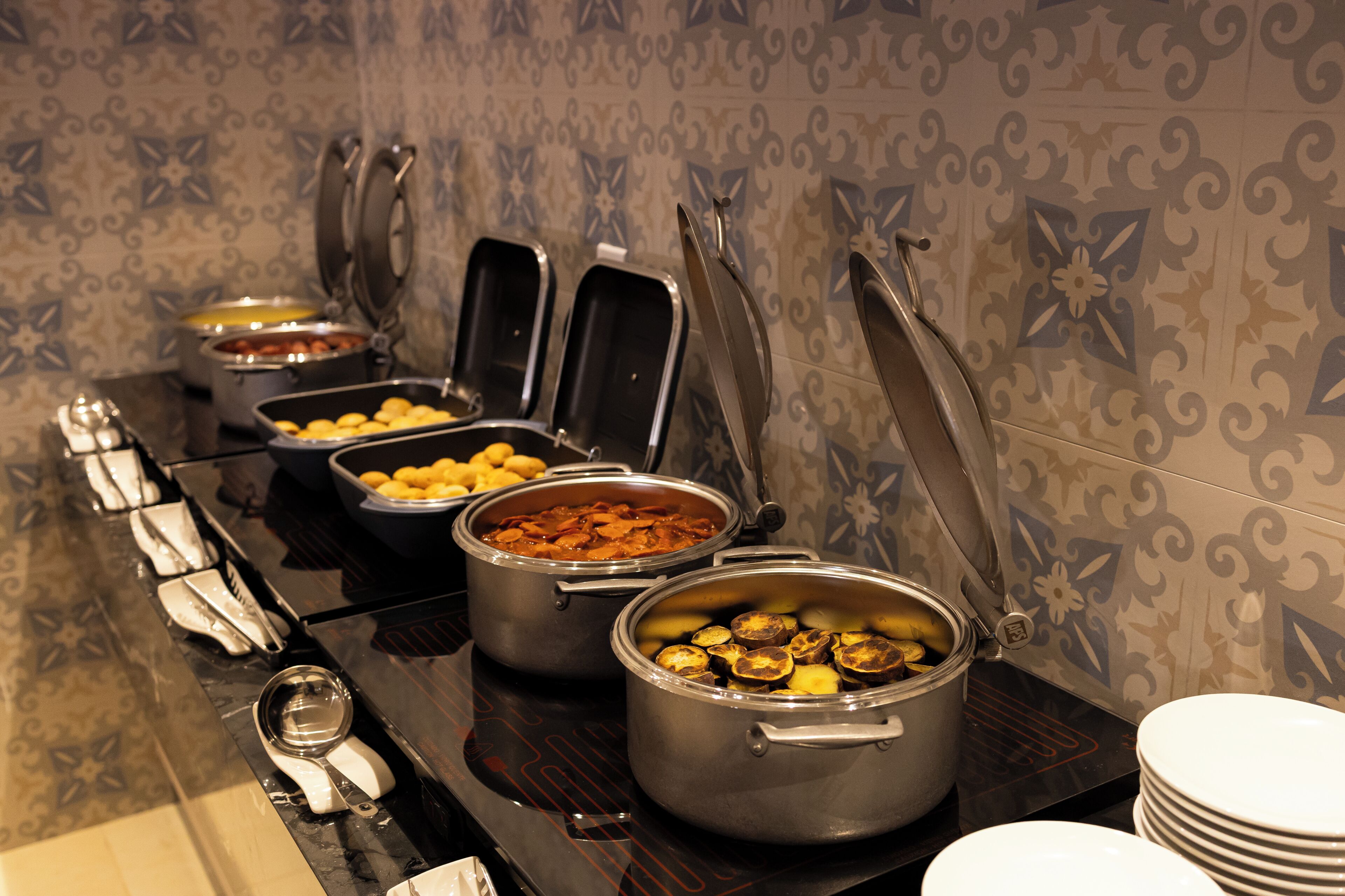 Una mesa de buffet con diversos alimentos en ollas y recipientes, una pila de platos y una cuchara.