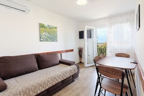 Ein Zimmer mit einer Couch, einem Esstisch mit Stühlen, einem an der Wand montierten Fernseher und einer Tür zu einem Außenbereich.