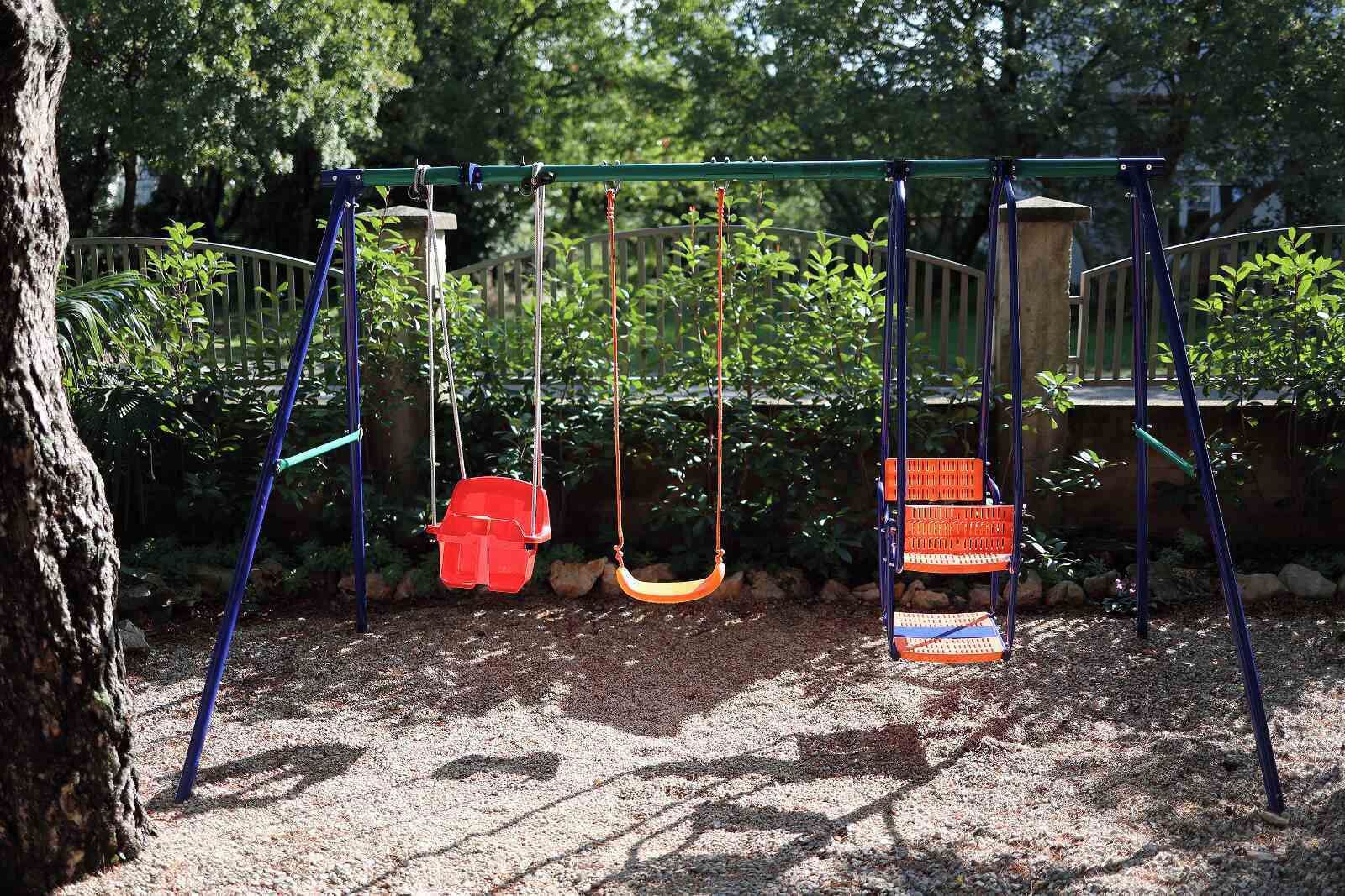 Eine Schaukel in einem Park, im Hintergrund ein Baum und eine hölzerne Konstruktion.