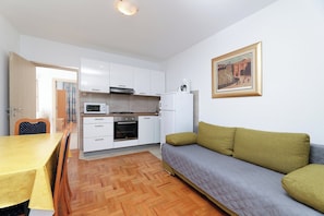 Eine kompakte Küche mit weißen Schränken, einer Mikrowelle, einem Backofen und einem Kühlschrank. Ein Esstisch mit Stühlen und ein Sofa mit Kissen sind vorhanden. Ein Gemälde hängt an der Wand.