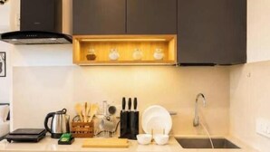 A modern kitchen with dark cabinets, a wooden shelf, and a sink. - Olyra Living | Cozy Luxury Near Airport (MH)