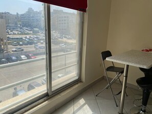 Een kamer met een groot raam, een eettafel met twee stoelen en uitzicht op een parkeerplaats en gebouwen.