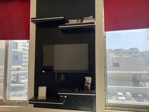 Een kamer met een groot raam, een zwarte, aan de muur bevestigde televisie, een klein plankje met boeken en een rood rolgordijn.