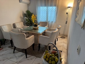 A dining area with a glass table surrounded by white chairs, a potted plant, and a basket of fruits.