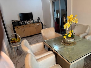 Un comedor con una mesa de vidrio, sillas color beige, una cesta de frutas y una cocina al fondo.