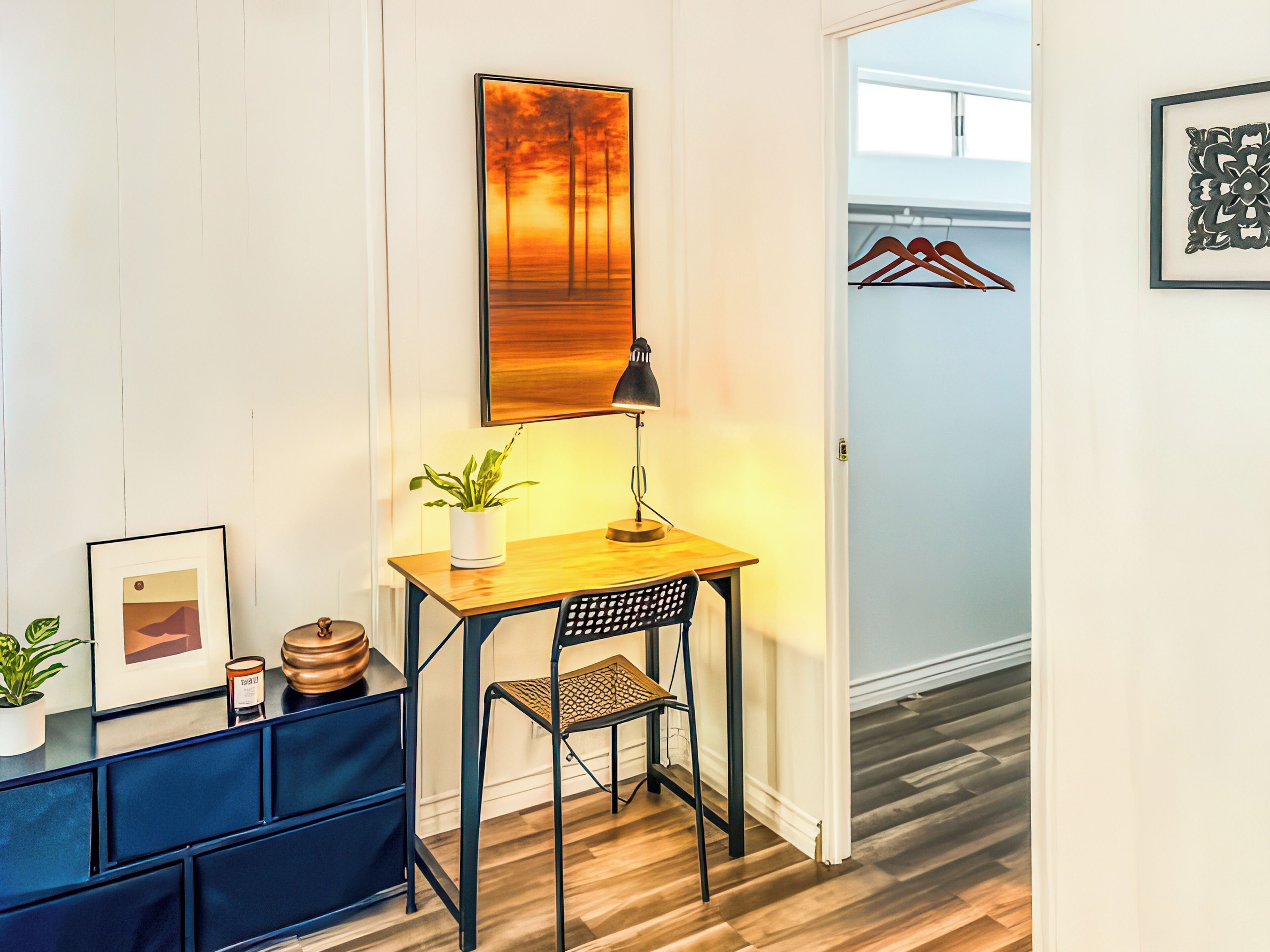 A small room with a desk, chair, lamp, potted plant, and framed picture.