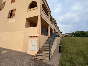 A building with a staircase leading to an upper level, a small door, and a grassy area beside it.