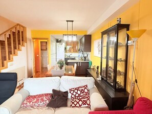 Una sala de estar con un sofá, un comedor con mesa y sillas, una cocina al fondo y una escalera.