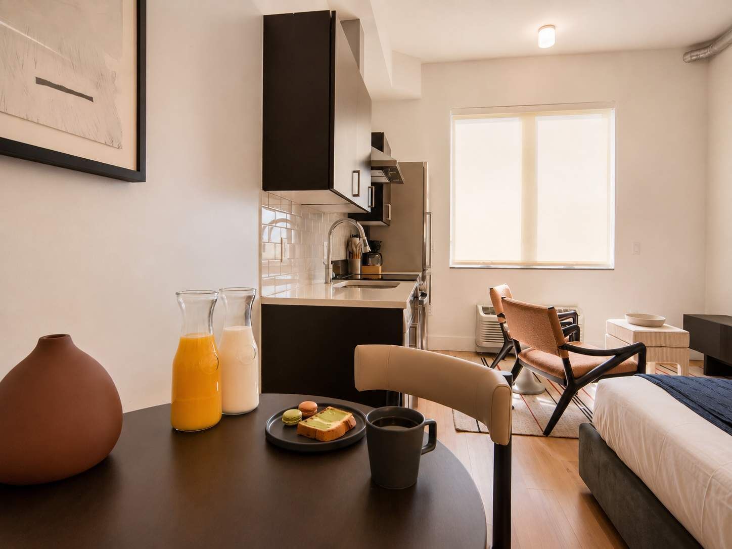 A modern hotel room with a dining area, kitchenette, and a large window providing natural light.