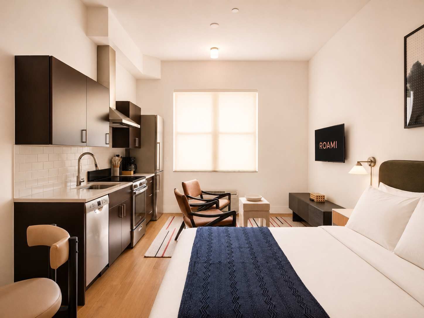 Una habitación de hotel moderna con kitchenette, una cama con un plaid azul, un televisor y una ventana.
