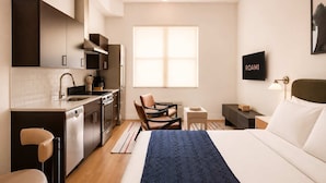A modern hotel room with a kitchenette, a bed with a blue throw, a TV, and a window. - Roami at Studio 135 - Modern Studio (Denver)