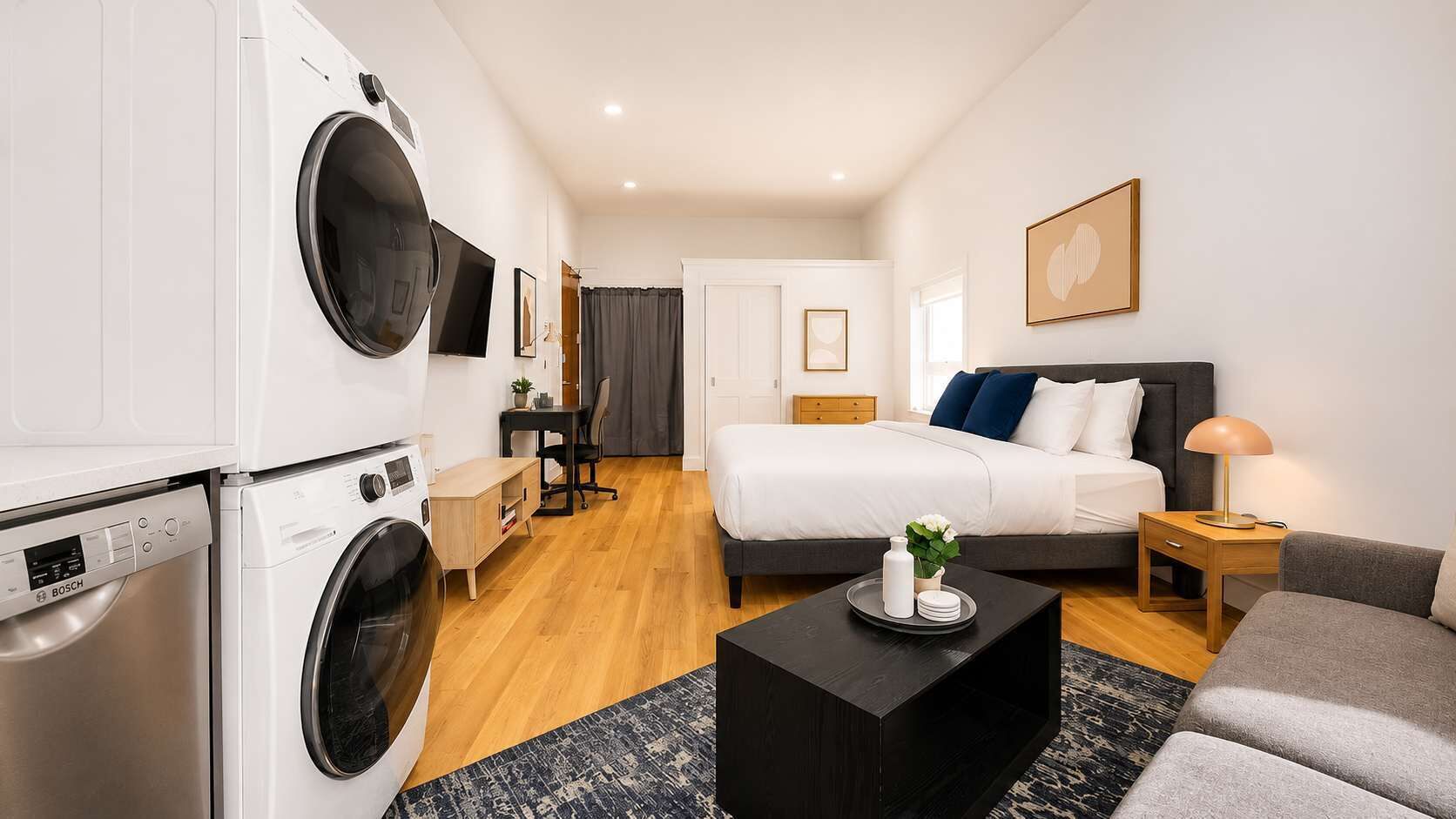 A modern hotel room with a washing machine, a bed, a desk, and a sofa.