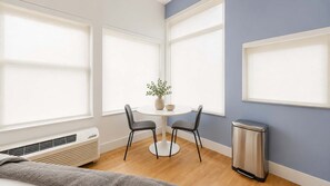A room with a round table, two chairs, a potted plant, a trash bin, and a window with blinds. - Roami at Studio 135 - Deluxe Studio (Denver)