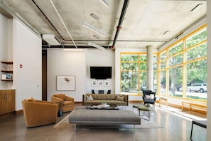 Un interior moderno con un sofá, una otomana y sillones, un televisor de pantalla plana y ventanas amplias con vistas a árboles y coches aparcados.
