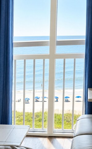 Una habitación con un ventanal grande que da a una playa con sombrillas y una barandilla blanca.