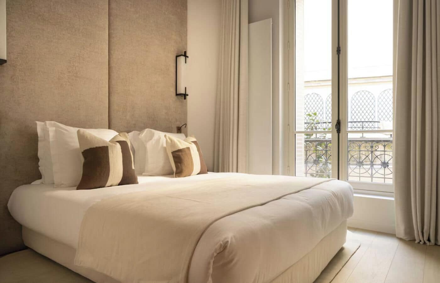 Ein großes Bett mit neutraler Farbgestaltung, eine Wandleuchte und Blick auf einen Balkon mit Metallgeländern.