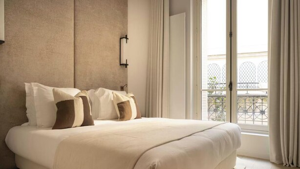 Una cama grande con una paleta de colores neutros, un aplique de pared y vistas a un balcón con barandillas de metal.