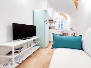 Ein modernes Wohnzimmer mit einem Flachbildfernseher auf einem weißen Schrank, einem türkisblauen Kissen auf einer weißen Couch und einer kleinen Küchenzeile im Hintergrund.