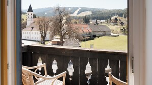 A balcony with wicker chairs, a view of a town with a church and a building with a red roof, and a tree in the foreground. - Aparthotel Hirschenhof (Spital am Semmering)