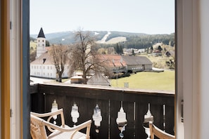 Un balcon avec des fauteuils en osier, une vue sur une ville avec une église et un bùtiment à toit rouge, et un arbre au premier plan.