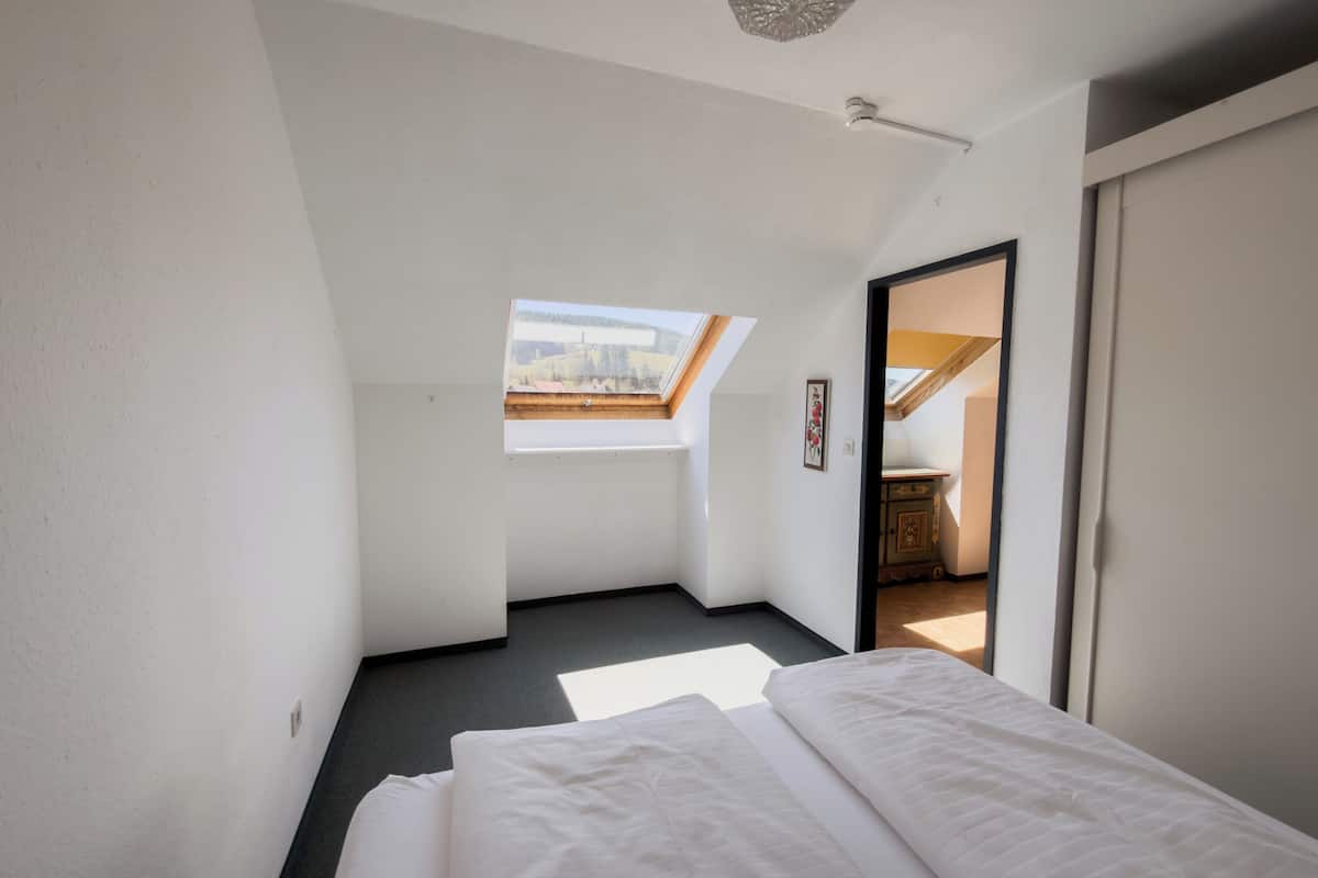 A bedroom with a skylight, a bed, and a view of a room with a wooden ceiling.